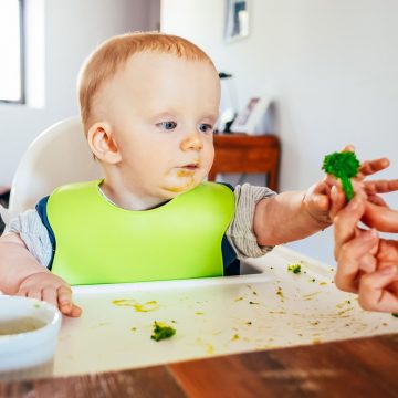 Baby im Hochstuhl, dass nach einem Stück Broccoli greift