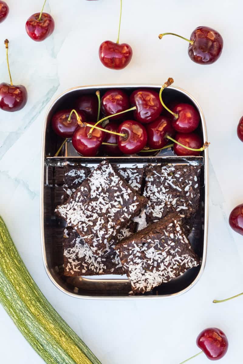 Zucchini-Brownies, mit Kokosraspeln bestreut, in einer Brotdose, mit Kirschen