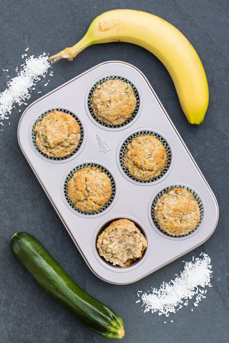 Zucchini-Muffins in einer Muffinform, daneben liegt eine Zucchini, eine Banane und Kokosraspeln