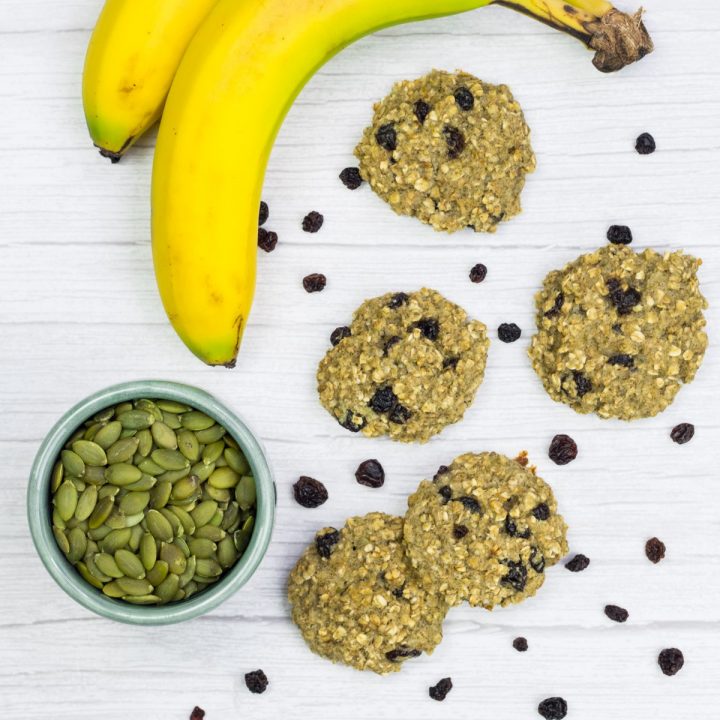 Haferflockenkekse ausgebreitet, daneben liegen Kürbiskerne, Rosinen und eine Banane