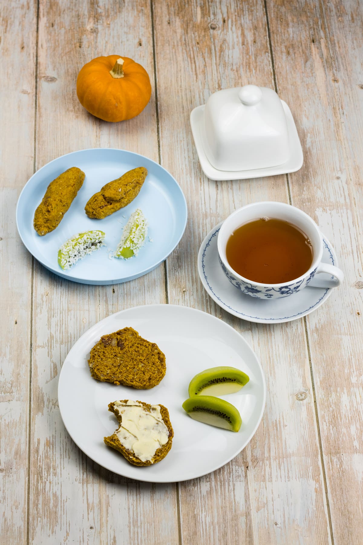 Veganer Scone aufgeschnitten auf einem Teller und mit Butter bestrichen, daneben eine Tasse Tee und ein Babyteller mit länglichen Scones und Kiwi, ein einfaches Kürbis Rezept