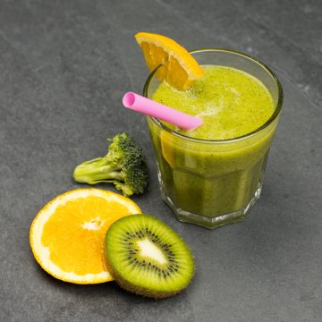 Vitamin C Smoothie in einem Glas, daneben eine Scheibe Orange, Kiwi und ein Stück Brokkoli