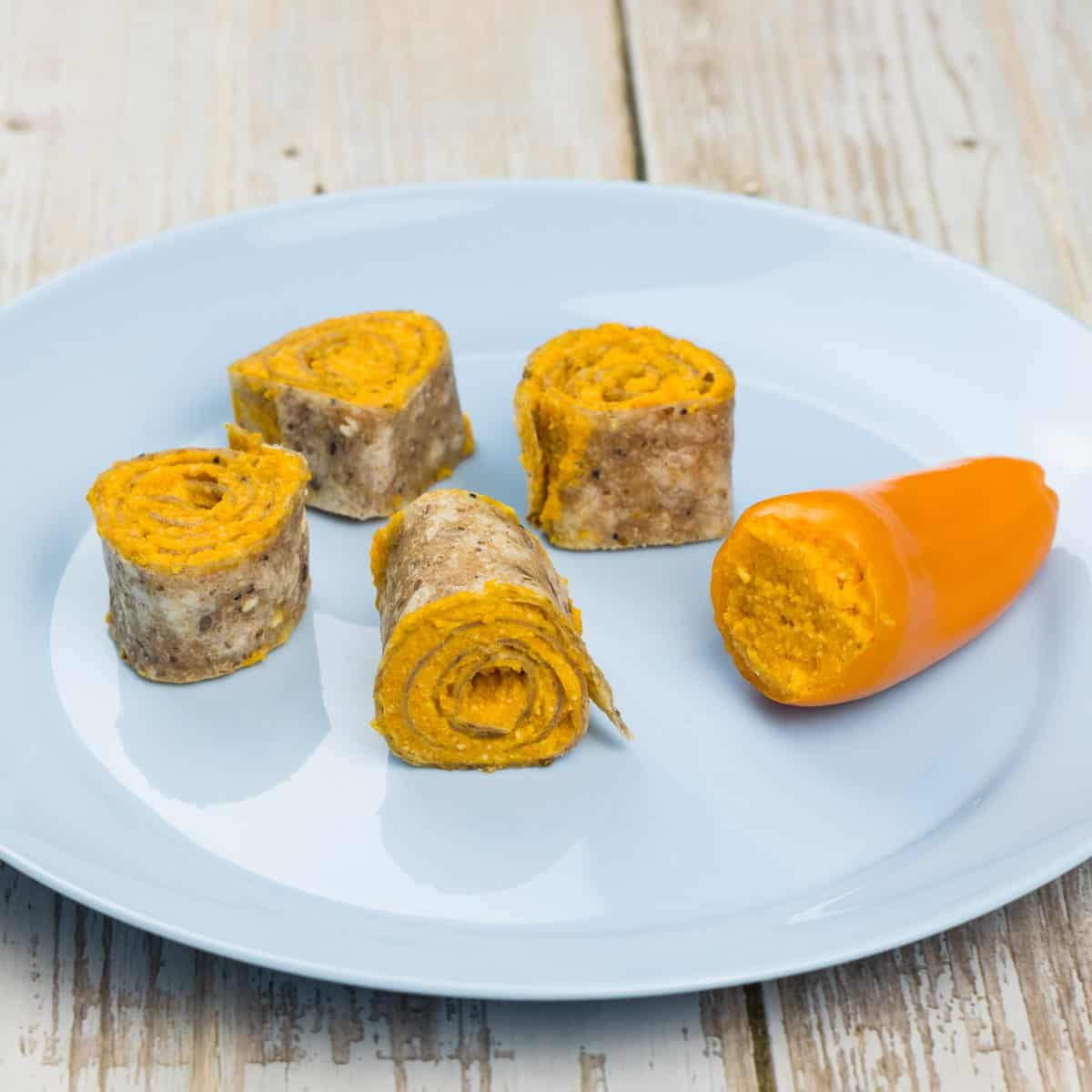 Ein Wrap mit Karottenaufstrich bestrichen, aufgerollt und in Scheiben geschnitten, daneben liegt eine Mini-Paprika, die mit Karottenaufstrich gefüllt ist