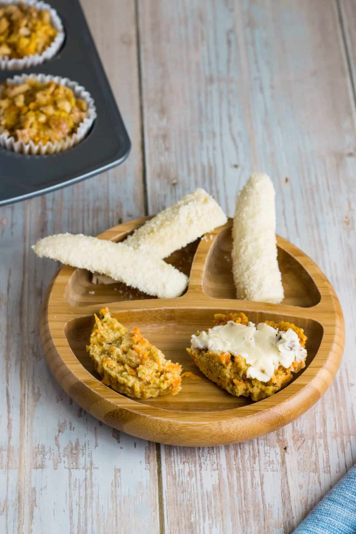 Karottenmuffins ohne Zucker für Babys, auf einem Babyteller, mit Butter beschmiert, daneben ein Stück Banane