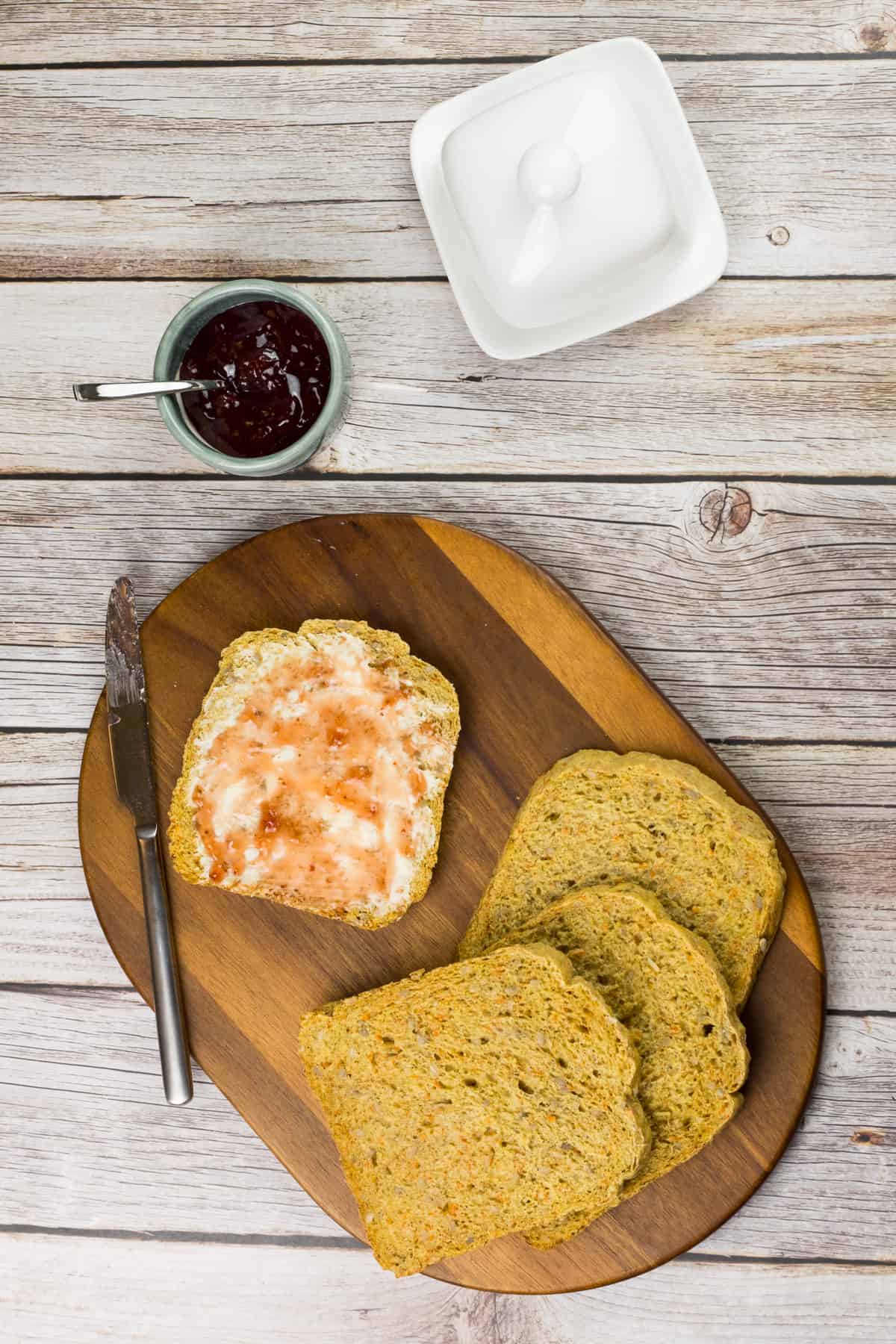 Karottenbrot mit Sonnenblumenkernen, aufgeschnitten auf einem Holzbrett, eine Scheibe ist mit Butter und Marmelade beschmiert