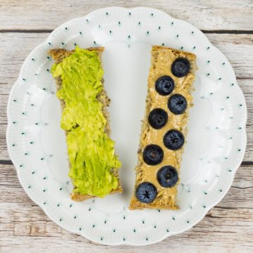 Zwei längliche Stücke Brot für ein Baby, auf einem Teller, ein Stück ist mit Avocadomus bestrichen, das andere mit Nussmus und halbierten Blaubeeren