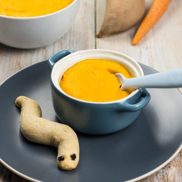 Süßkartoffel-Möhren-Suppe in einer Schale für Kinder, daneben liegt ein Wurm aus Brot zum Dippen