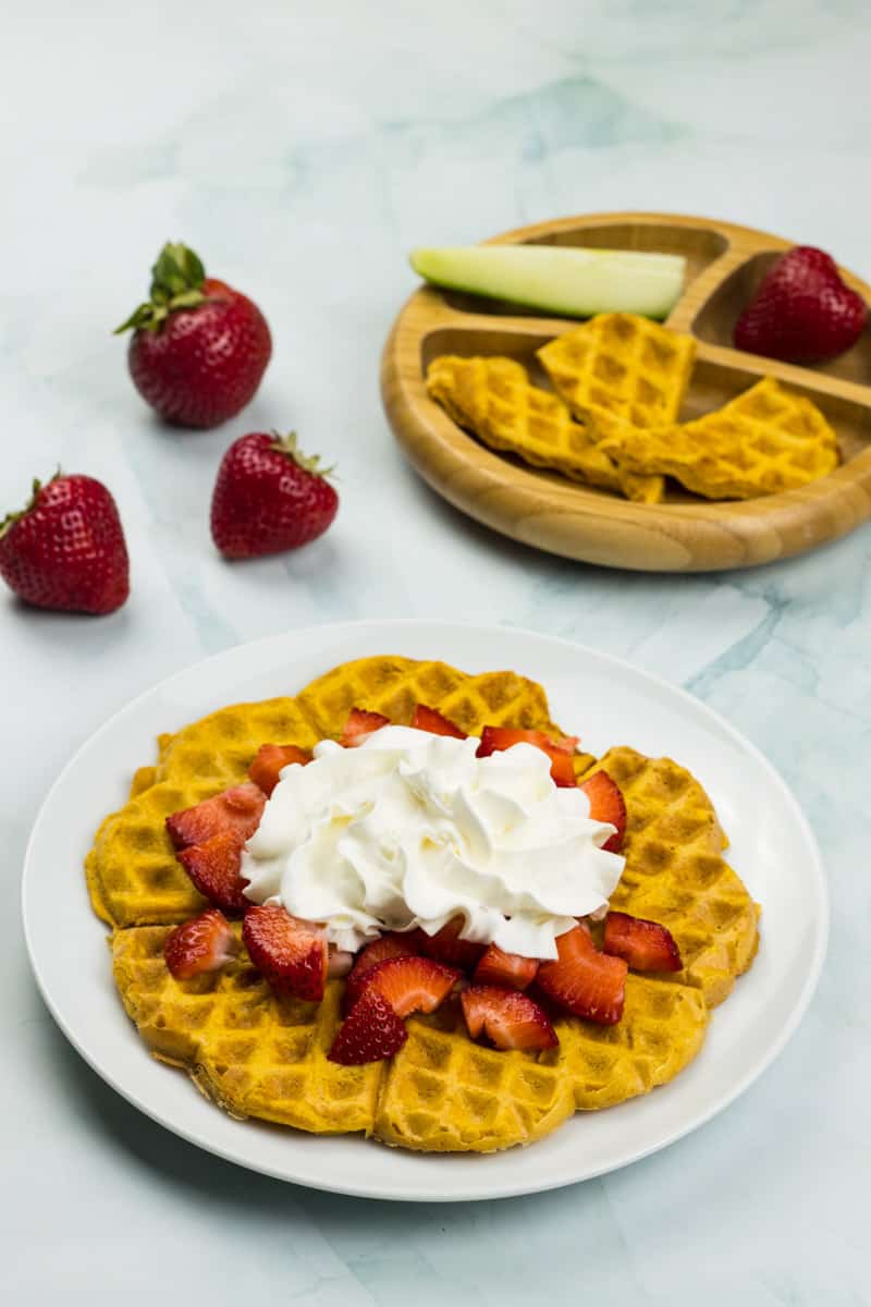 Süßkartoffel Waffel auf einem Teller, mit Erdbeeren und Schlagsahne belegt, dahiner ein Babyteller mit Süßkartoffel Waffeln