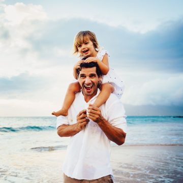 Kleines Mädchen sitzt auf dem Schultern vom Vater, am Strand