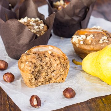 ein halber Birnen-Muffin, auf einem Holzbrett, dahinter liegen mehr Birnenmuffins, Birnen und Haselnüsse