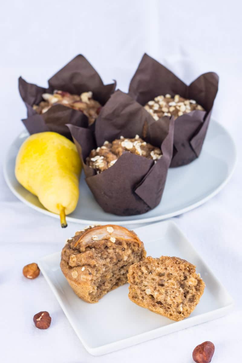 Gesunde Birnen-Muffins ohne Zucker, auf einem Teller, in zwei Hälften gebrochen, dahinter steht noch ein Teller mit Birnenmuffins, einer Birne und Haselnüssen