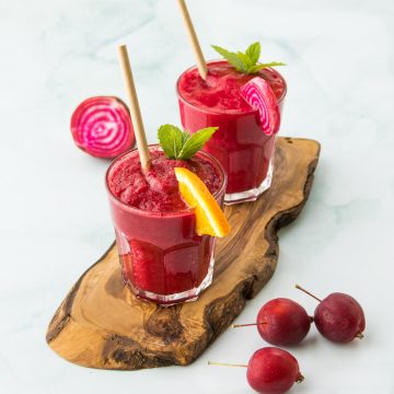 Zwei Gläser mit Rote Bete Smoothie, mit Orange und Minze dekoriert, auf einem Holzbrett, daneben Äpfel und Rote Beete