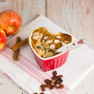 Apfel-Grieß-Auflauf in einer Herz-Schüssel, mit Babylöffel, daneben liegen Äpfel, Rosinen und Zimtstangen