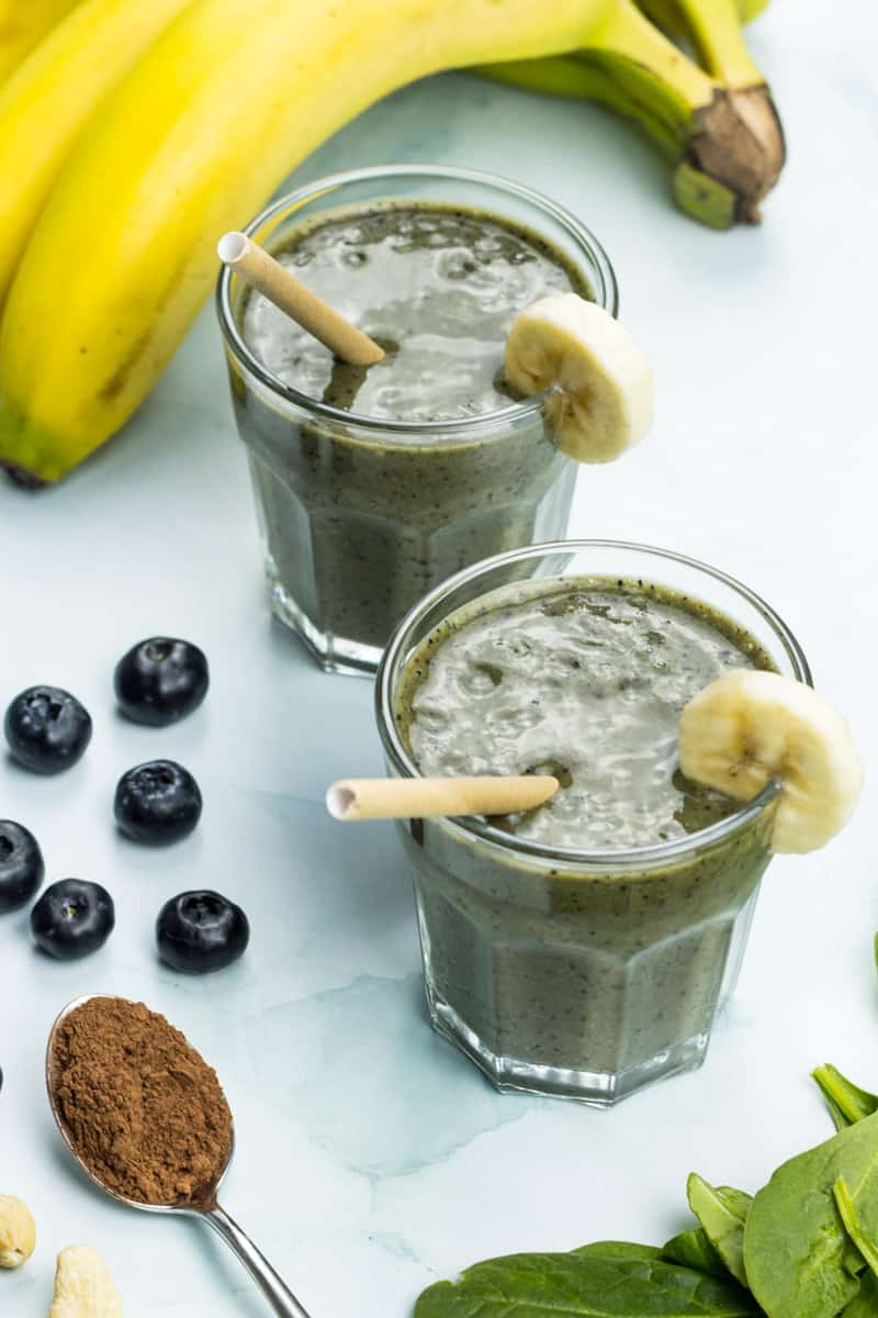 zwei Gläser mit gesundem, veganen Banananshake mit viel Magnesium, daneben liegen Blaubeeren, Cashewnüsse, Spinat und Banane