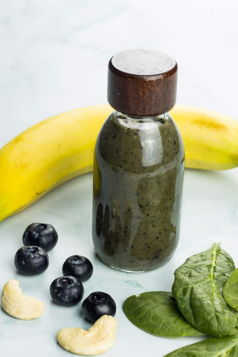 gesunder, veganer Banananshake mit viel Magnesium, in einer Smoothie Flasche, daneben liegen Blaubeeren, Cahewnüsse, Spinat und Banane