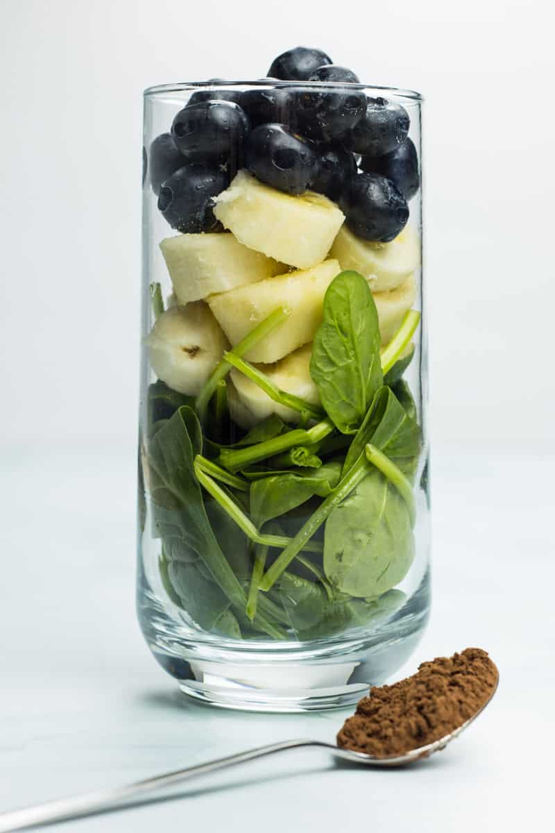 Glas gefüllt mit Spinat, Banane und Blaubeeren, mit einem Teelöffel Kakao davor, für einen veganen Bananenshake