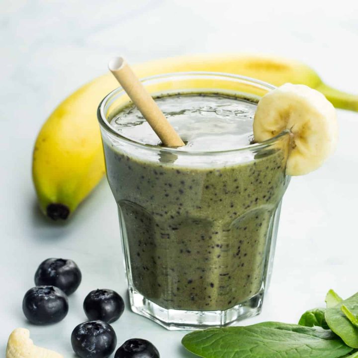 gesunder, veganer Bananenshake mit viel Magnesium, in einem Glas, daneben liegen Blaubeeren, Cashewnüsse, Spinat und Banane