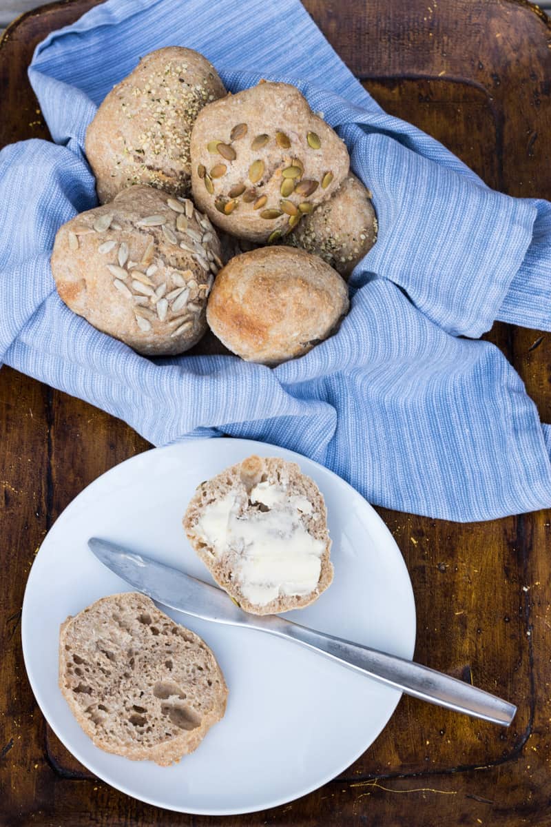 Einfache Dinkelbrötchen über Nacht, in einem Brotkorb, ein fluffiges Brötchen ist aufgeschnitten auf einem Teller, mit Butter beschmiert