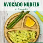 cremige Avocado-Nudeln auf einem Babyteller, mit grünem Spargel und einem Stück Orange