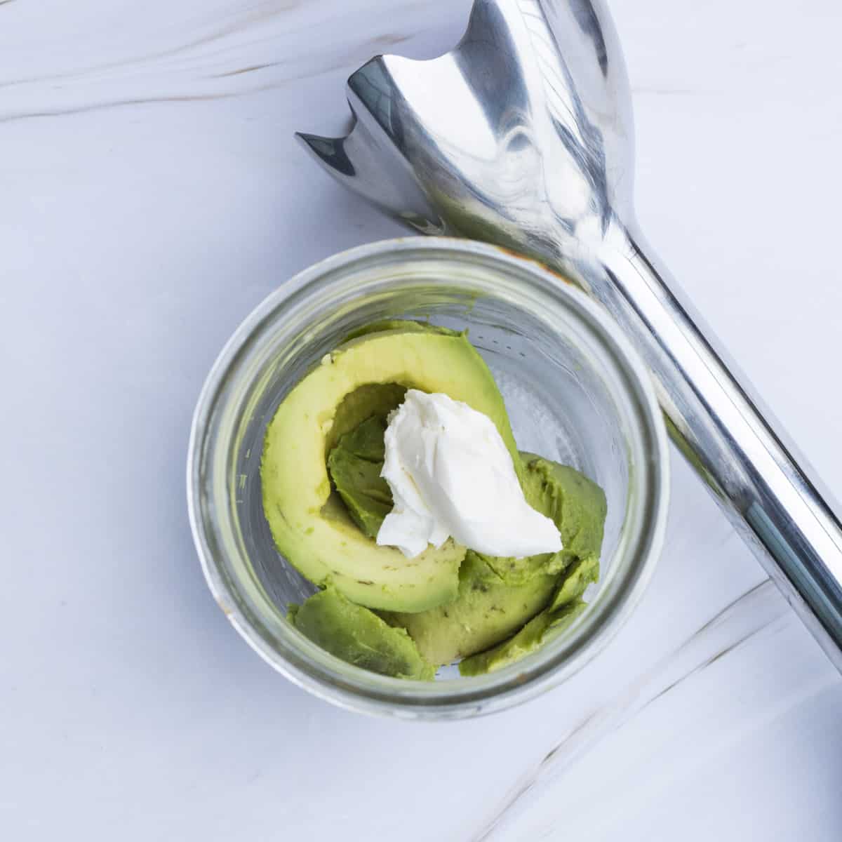 eine halbe Avocado und etwas Frischkäse in einem Glas, daneben ein Pürierstab zum Pürieren