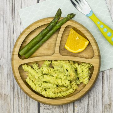 cremige Avocado-Nudeln auf einem Babyteller, mit grünem Spargel und einem Stück Orange