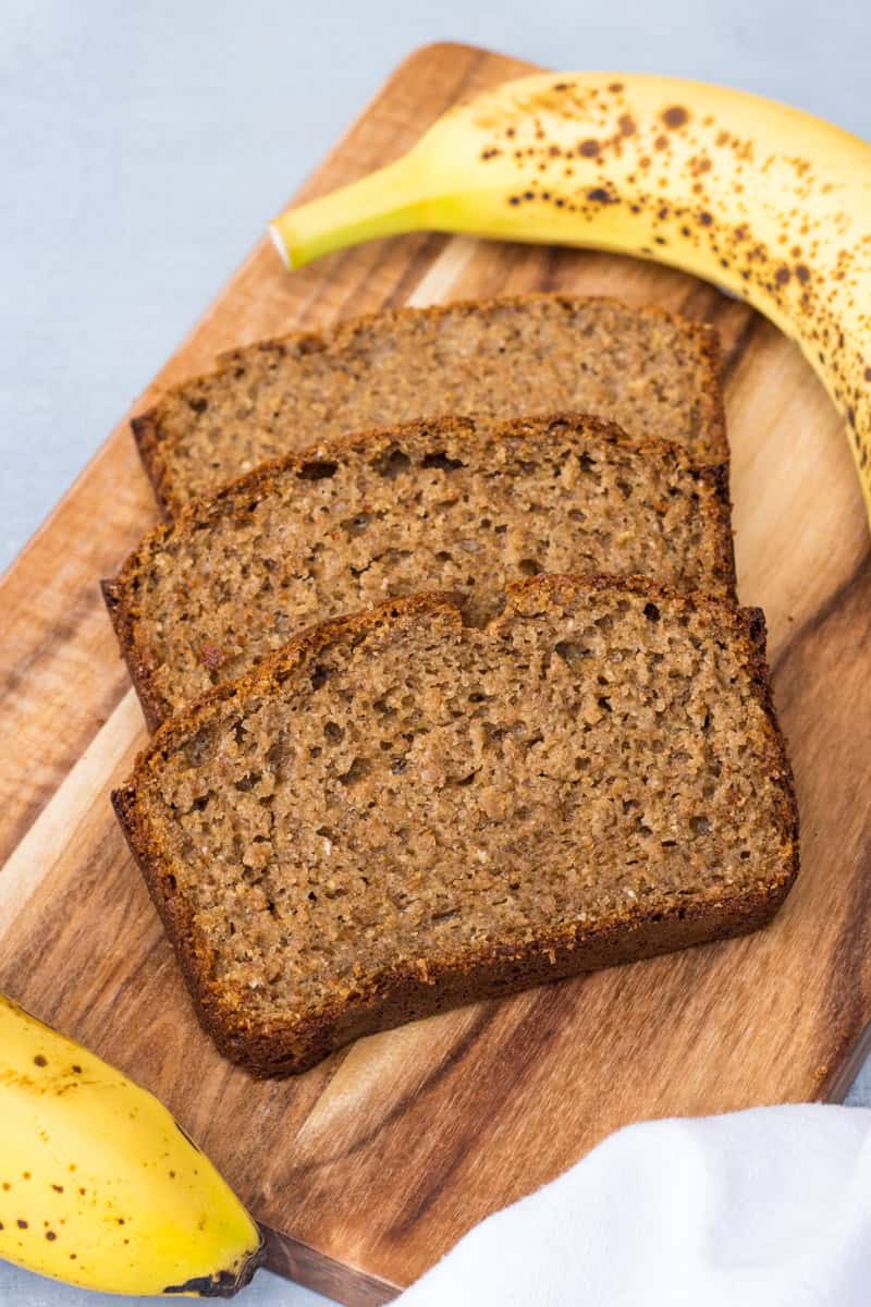 Drei Scheiben Bananenbrot auf einem Holzbrett, daneben liegen reife Bananen